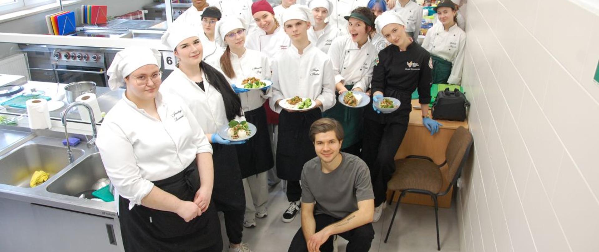 Młodzież w strojach kucharskich wokół prowadzącego warsztaty w pracowni gastronomicznej