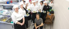 Młodzież w strojach kucharskich z przygotowanymi potrawami wokół prowadzącego warsztaty