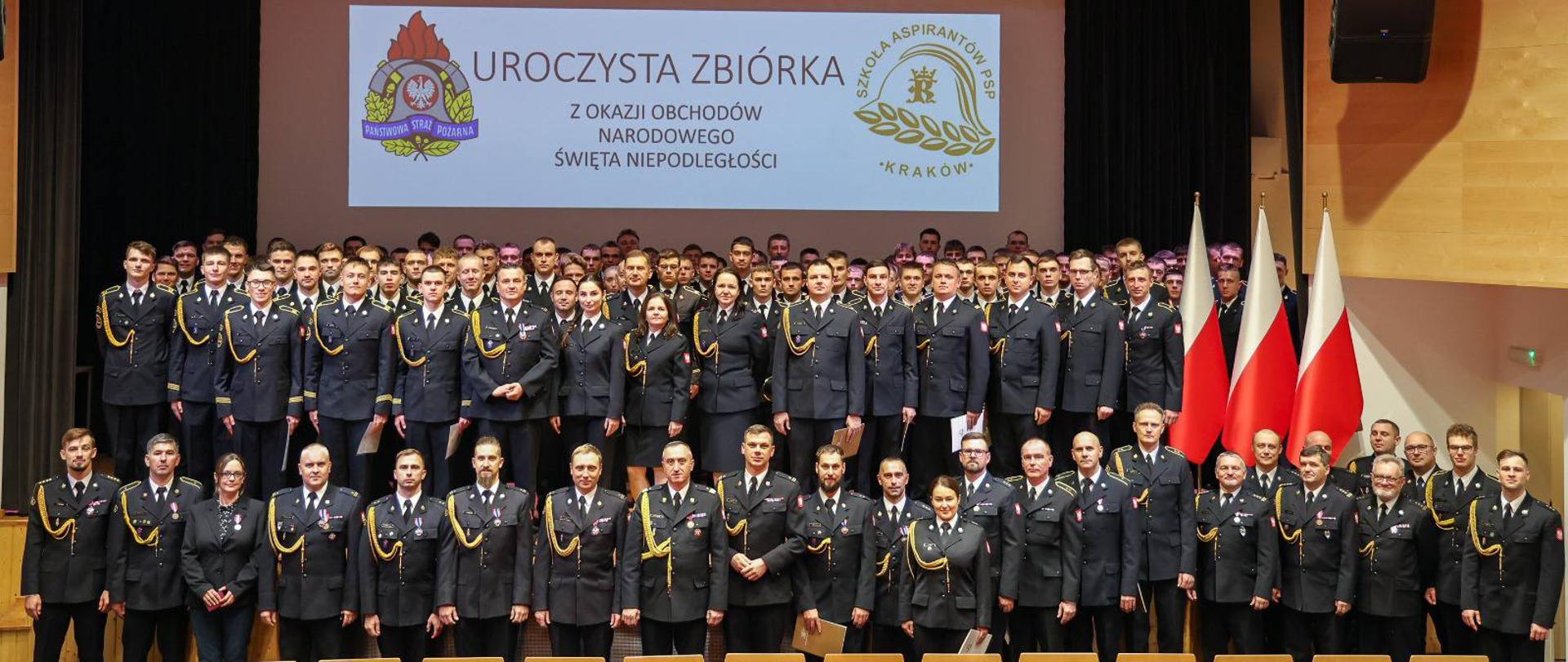 Zdjęcie dokumentuje uroczystą zbiórkę z okazji Narodowego Święta Niepodległości, która odbyła się w Szkole Aspirantów Państwowej Straży Pożarnej w Krakowie. Centralnym punktem kadru jest duża grupa, złożona z kadry dowódczej i kadetów, stojących w rzędach na scenie. Wszyscy uczestnicy ubrani są w galowe, ciemnogranatowe mundury PSP ze złotymi akcentami, a osoby z pierwszego rzędu, kadra, mają na piersiach przypięte medale i odznaczenia. W tle, na dużym ekranie, widnieje napis: "UROCZYSTA ZBIÓRKA Z OKAZJI OBCHODÓW NARODOWEGO ŚWIĘTA NIEPODLEGŁOŚCI", otoczony symbolami PSP i krakowskiej Szkoły Aspirantów. Po prawej stronie sceny, dumnie eksponowane, stoją polskie flagi państwowe, podkreślając patriotyczny charakter uroczystości, podczas której Komendant wręczał odznaczenia i awanse, nawiązując do wartości wolności i służby.