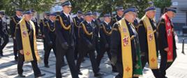 Funkcjonariusze PSP w mundurach galowych z odznaczeniami i sznurami galowymi maszerują aleją w kierunku Jasnej Góry. Wśród nich widoczni są trzej kapelani Straży Pożarnej w żółtej stule liturgicznej oraz jeden w czerwonej stule liturgicznej