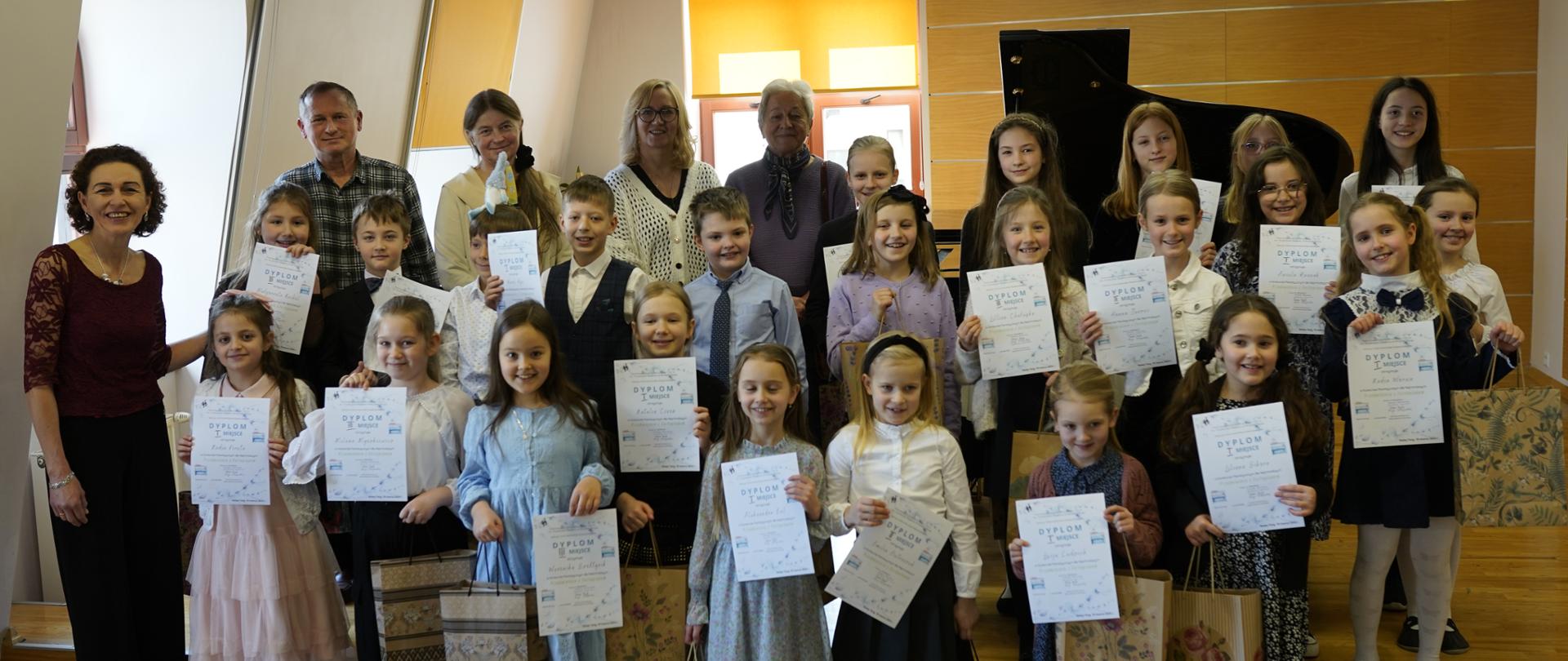 Wszyscy uczestnicy konkursu wraz z jurorami i prowadzącą konkurs. Dzieci trzymają w dłoniach dyplomy laureatów a w drugiej torby papierowe z upominkami. Za nimi fortepian, trzy okna przysłonięte roletami.