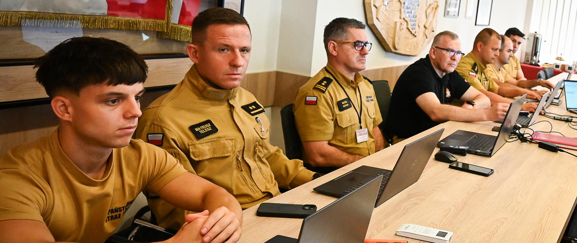 Grupa mężczyzn uczestniczących w warsztatach w Komendzie Wojewódzkiej PSP w Rzeszowie. Uczestnicy siedzą w rzędzie przy długim, drewnianym stole, na którym rozstawione są otwarte laptopy, telefony i materiały do notowania. Pierwsze plan zajmują strażacy Państwowej Straży Pożarnej w piaskowych mundurach służbowych. W tle widać fragment sztandaru oraz ozdobny godło/płaskorzeźbę na ścianie.