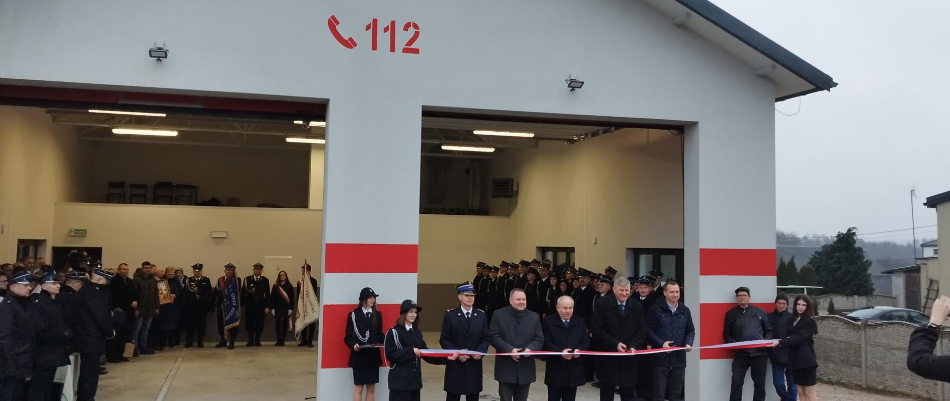 Zdjęcie przedstawia moment przecięcia wstęgi do strażnicy Ochotniczej Straży Pożarnej w Radoszkach. Po lewo zaproszeni goście zgromadzeni w remizie.