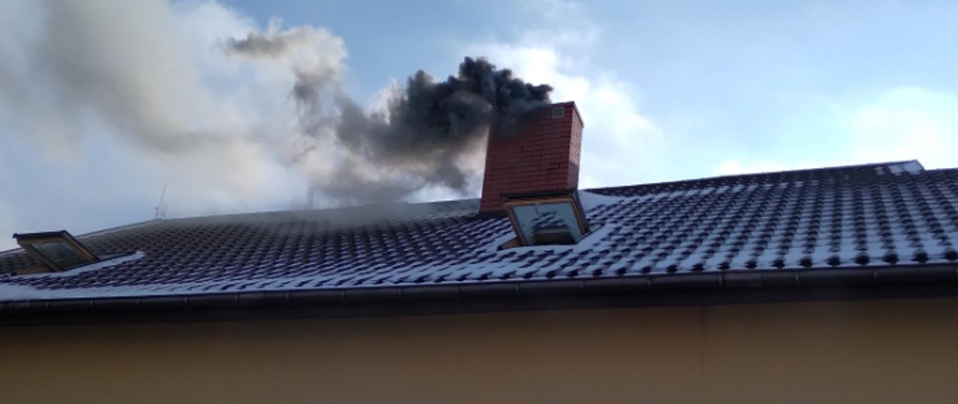 Zdjęcie przedstawia szary, gęsty dym wydobywający się z komina. Przed budynkiem w którym doszło do pożaru stoi dwóch strażaków.