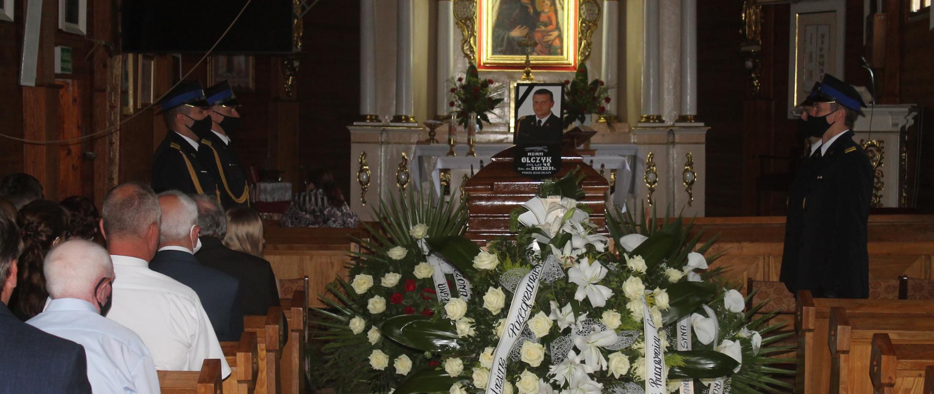 Zdjęcie przedstawia uroczystość pogrzebową st. ogn. Adama Olczyka we wnętrzu kościoła parafialnego w Umieniu. Na zdjęciu widać trumnę ze zdjęciem zmarłego, wartę honorową, wiązanki oraz zgromadzonych w kościele ludzi.