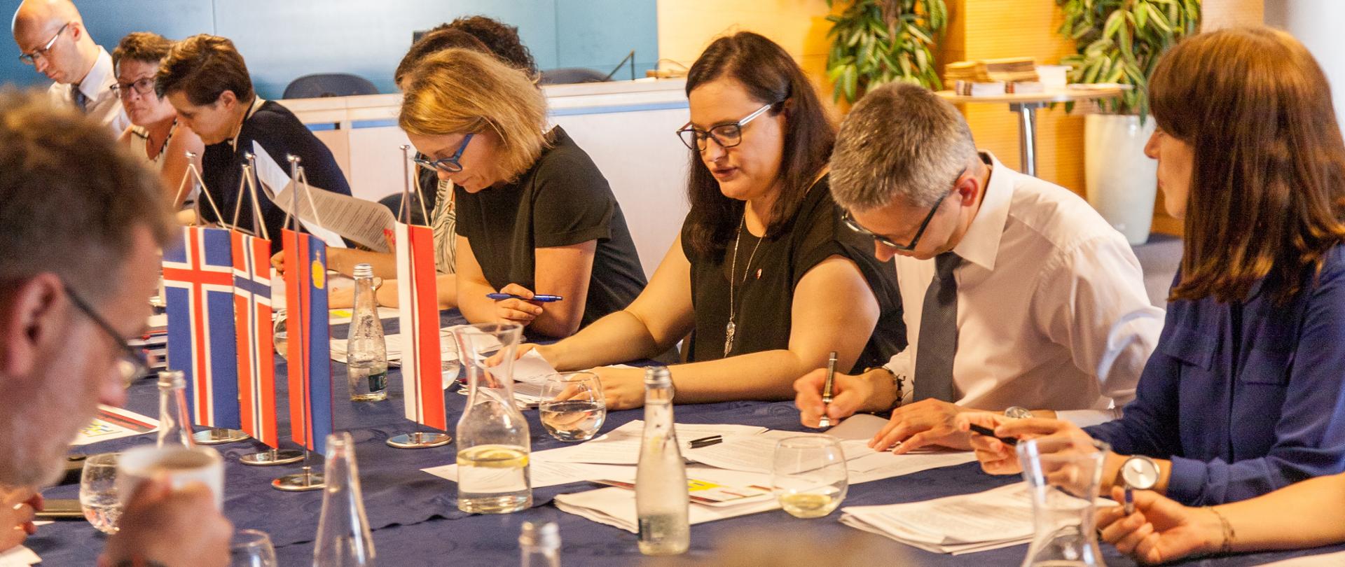 Na zdjęciu kilkanaście osób przy długim stole podczas obrad. Na stole proporczyki z flagami Polski i państw darczyńców - od lewej Islandii, Norwegii i Liechtensteinu. Trzecia z prawej wiceminister Małgorzata Jarosińska-Jedynak