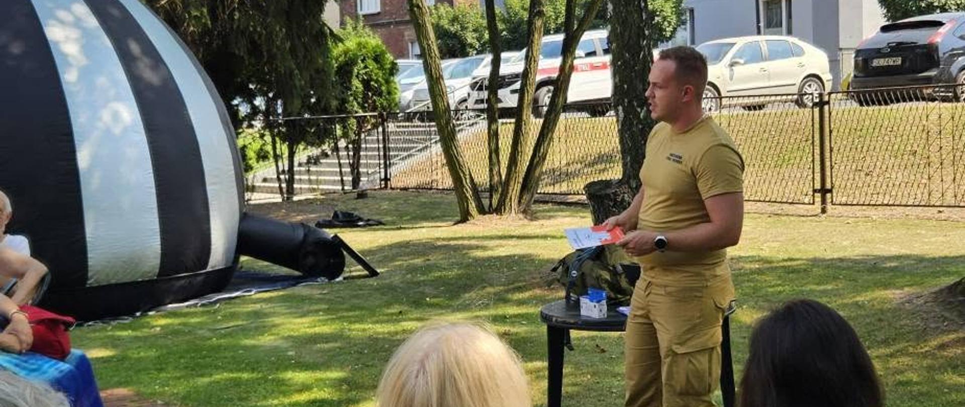 Na zdjęciu przedstawiono strażaka Państwowej Straży Pożarnej prowadzącego spotkanie edukacyjne dla grupy mieszkańców. Uczestnicy siedzą na zewnątrz, w otoczeniu zieleni, a w tle widoczny jest namiot pneumatyczny. Strażak podczas prelekcji omawia zasady bezpieczeństwa i właściwego postępowania w sytuacjach zagrożenia.