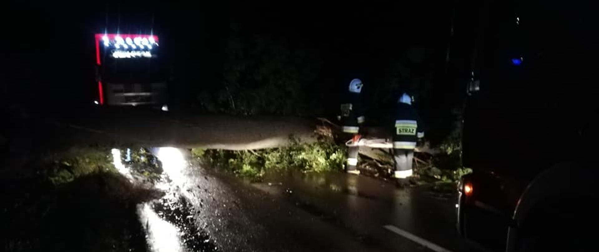 przewrócone drzewo blokuje drogę, strażacy przystępują do jego usuwania, w za drzewem stoi tir. 
