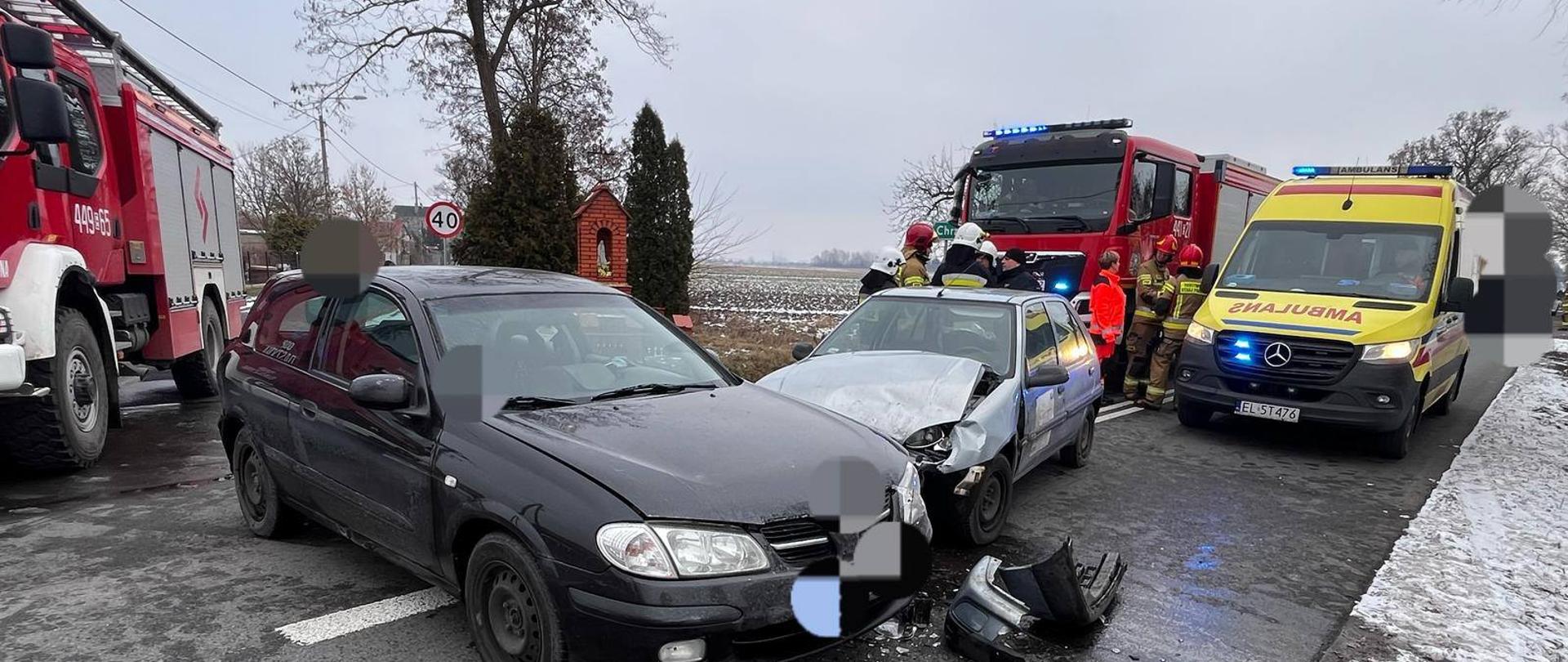 Na zdjęciu widać dwa rozbite samochody osobowe. Jeden pojazd jest koloru srebrnego a drugi czarnego. Oba stoją na jezdni. Za samochodami stoją dwa czerwone samochody straży pożarnej, żółta karetka pogotowia ratunkowego, strażacy oraz ratownik medyczny. W tle widać przydrożną kapliczkę, słup energetyczny, pole, znak drogowy z napisem 40, drzewa oraz krzewy. Jest widno.