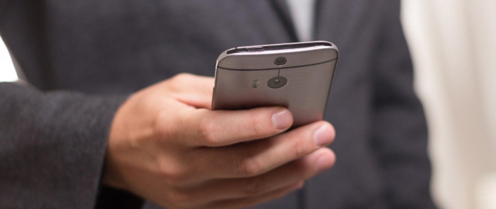 Mężczyzna w garniturze, w prawej dłoni trzyma smartfona.