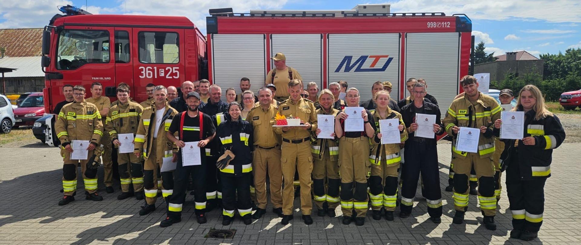 Zdjęcie grupowe strażaków którzy ukończyli szkolenie podstawowe. Strażacy stoją na tle pojazdu pożarniczego.