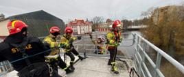 Trzech strażaków ubranych w hełmy specjalistyczne sprzęt wysokościowy trzymają niebieską linę obok stoi funkcjonariusz Państwowej Straży Pożarnej robi zabezpieczenie przed nimi strażak wspina się w zabezpieczeniu.