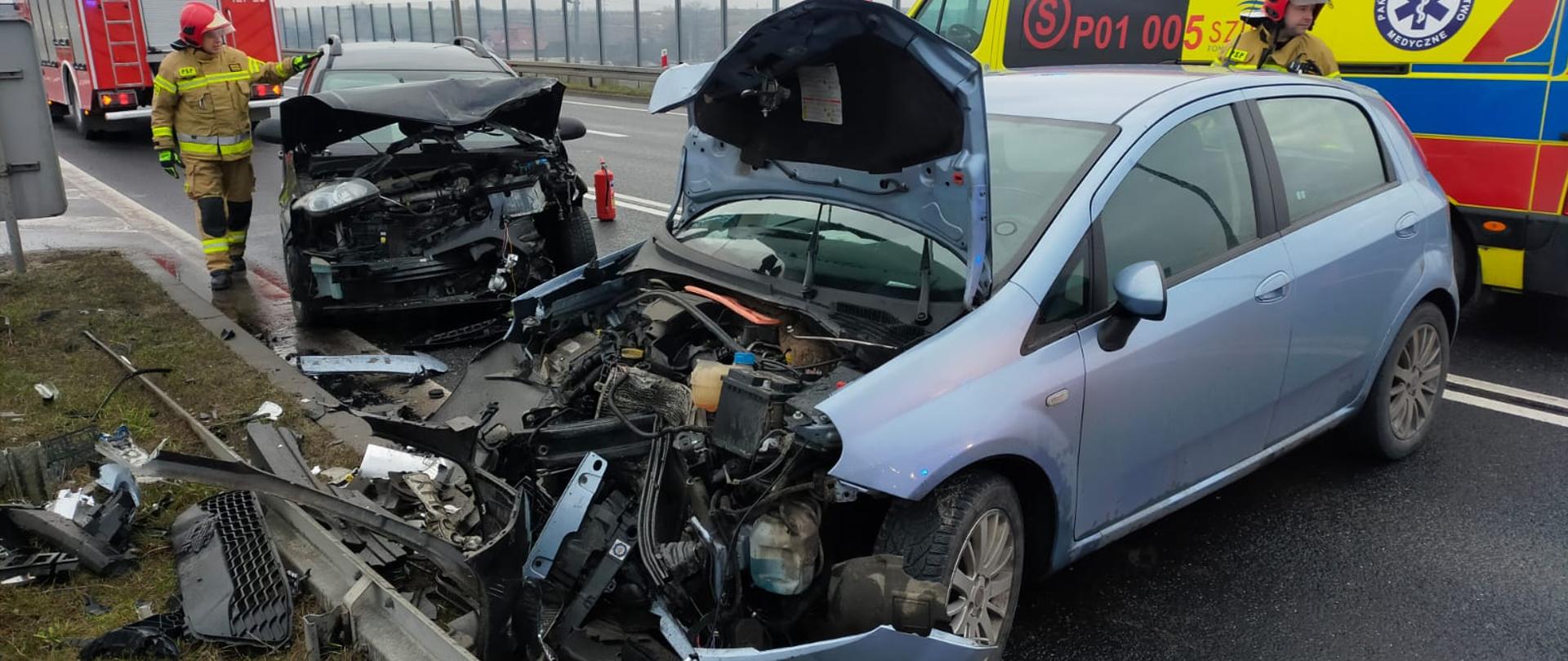 Na zdjęciu widać dwa pojazdy z uszkodzonymi przodami. W tle stoi samochód pożarniczy oraz karetka
