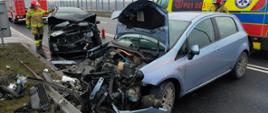 Na zdjęciu widać dwa pojazdy z uszkodzonymi przodami. W tle stoi samochód pożarniczy oraz karetka