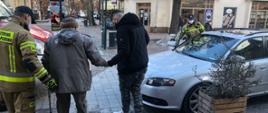 Zdjęcie przedstawia jak strażacy psp pomagają starszemu panu, z trudnościami w chodzeniu przejść z mieszkania do samochodu