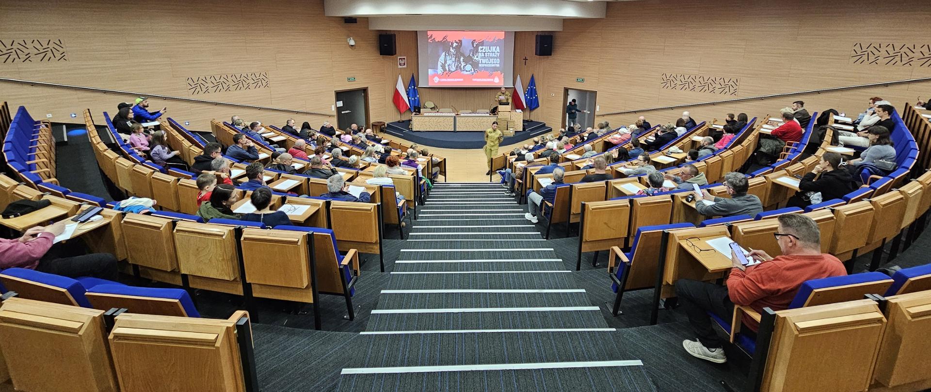 Widok z góry na siedzących w auli Centralnej Szkoły PSP osób przybyłych na spotkanie, w głębi pośrodku podium z mównic a przy której przemawia strażak. Za nim na ekranie multimedialnym wyświetlony jest slajdem prezentacji