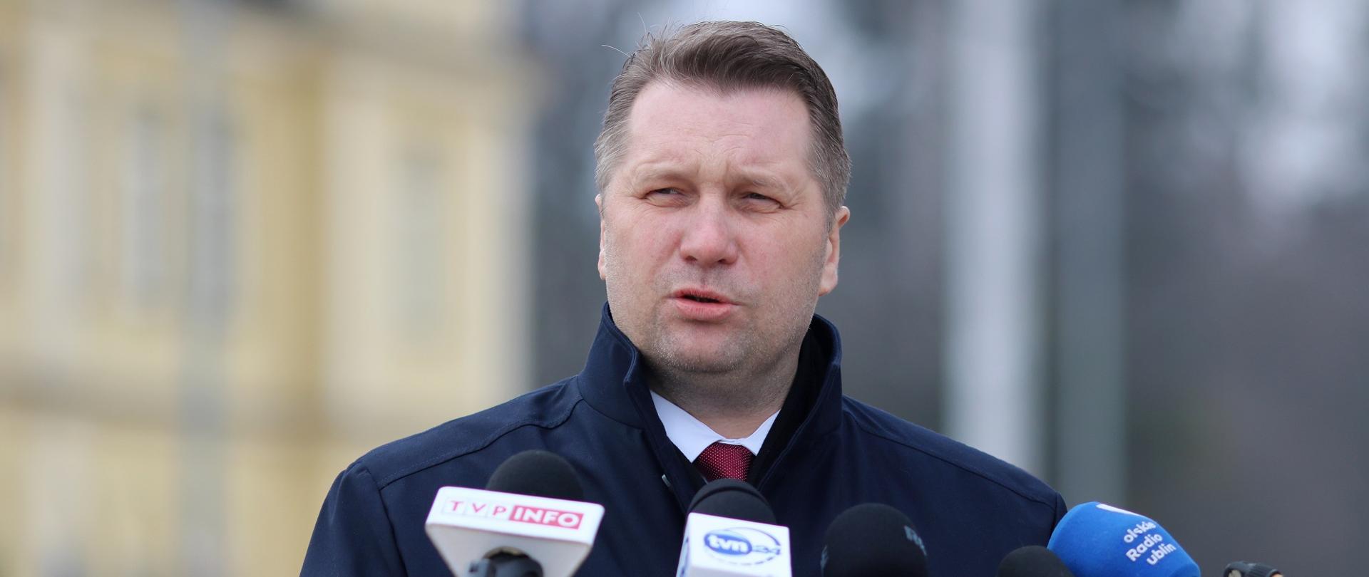 Minister Czarnek stoi i mówi do kilku mikrofonów, za nim w tle zarys budynku stacji kolejowej.