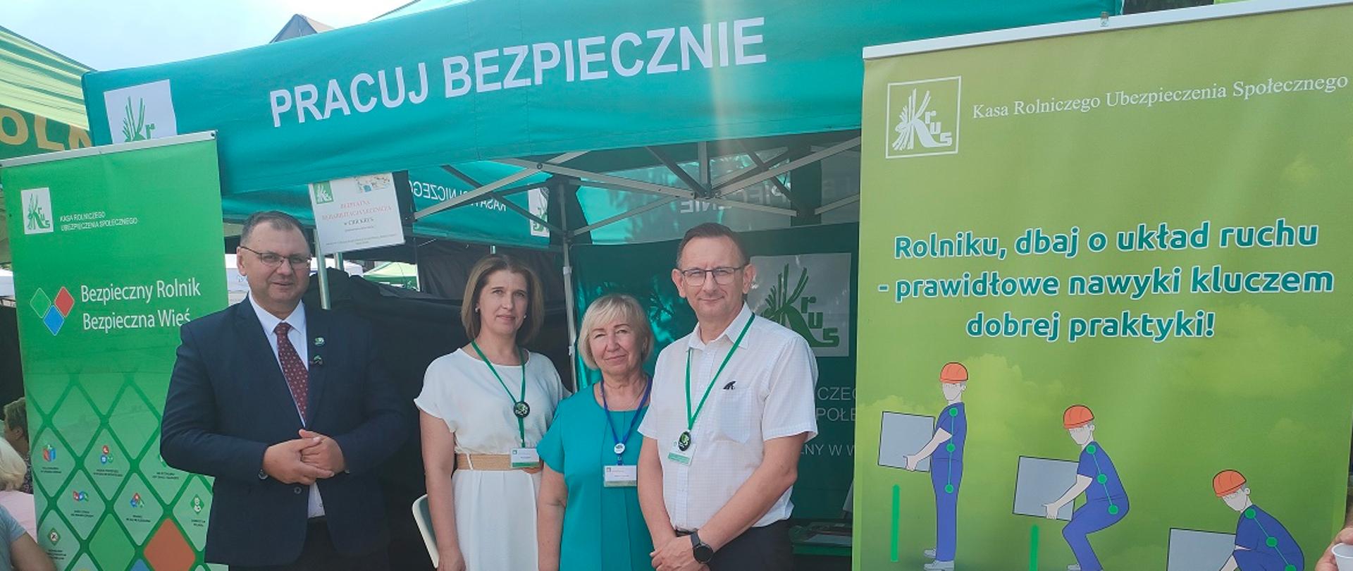 Grupa czterech osób dorosłych pozuje do zdjęcia przed zielonym namiotem informacyjnym KRUS, (dwie kobiety i dwóch mężczyzna) podczas wydarzenia plenerowego. Po bokach stoją zielone banery z logiem KRUS, na tym z prawej jest napis” „Rolniku, dbaj o układ ruchu – prawidłowe nawyki kluczem dobrej praktyki!”, na tym z lewej widnieje napis „Bezpieczny Rolnik Bezpieczna Wieś”. 