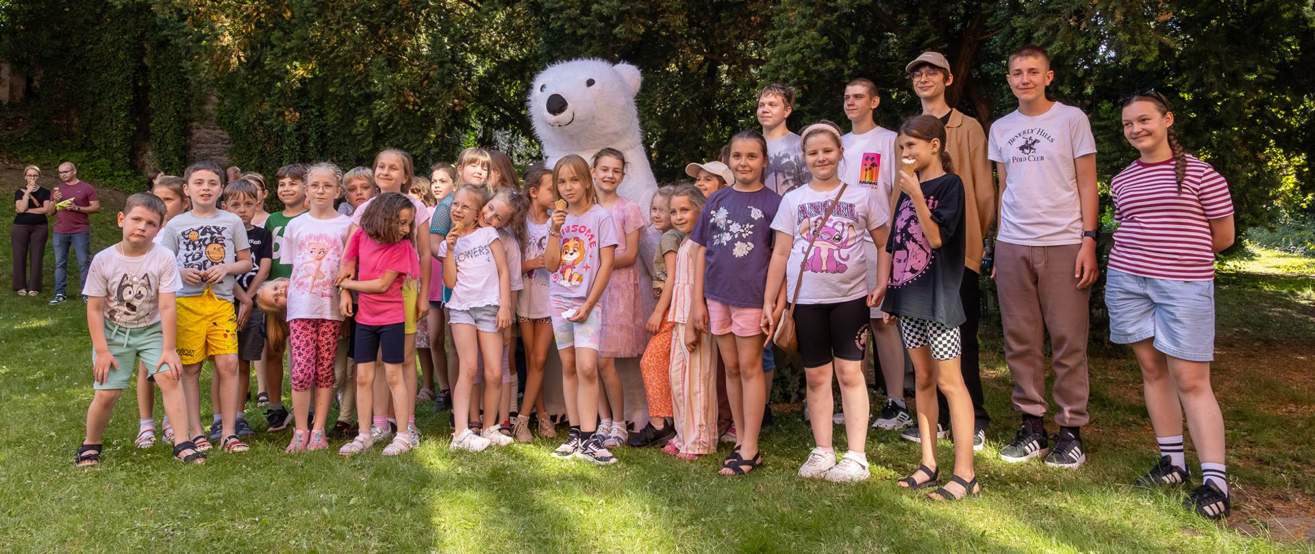 Uczestnicy pikniku rodzinnego, głównie dzieci, na wspólnej fotografii z misiem polarnym - maskotką animatorem zaproszonym na piknik.
