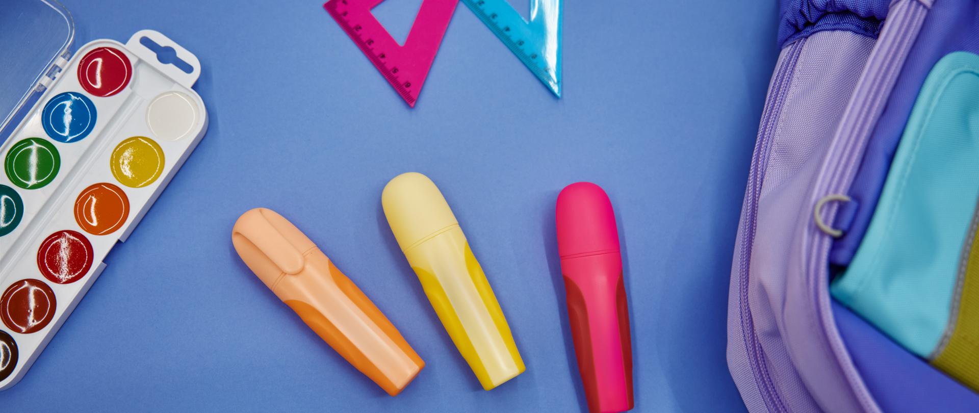 Backpack with watercolor paints, markers and ballpoint pens colorful school supplies on blue background. Back to school flat lay copy space