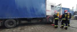 Na zdjęciu widać dym wydobywający się z kabiny samochodu ciężarowego oraz strażaków ubranych w ubrania bojowe i aparaty ochrony dróg oddechowych gaszących pożar w kabinie samochodu. Obok samochodu stoją również pracownicy. W tle widać inną ciężarówkę oraz przyczepy.