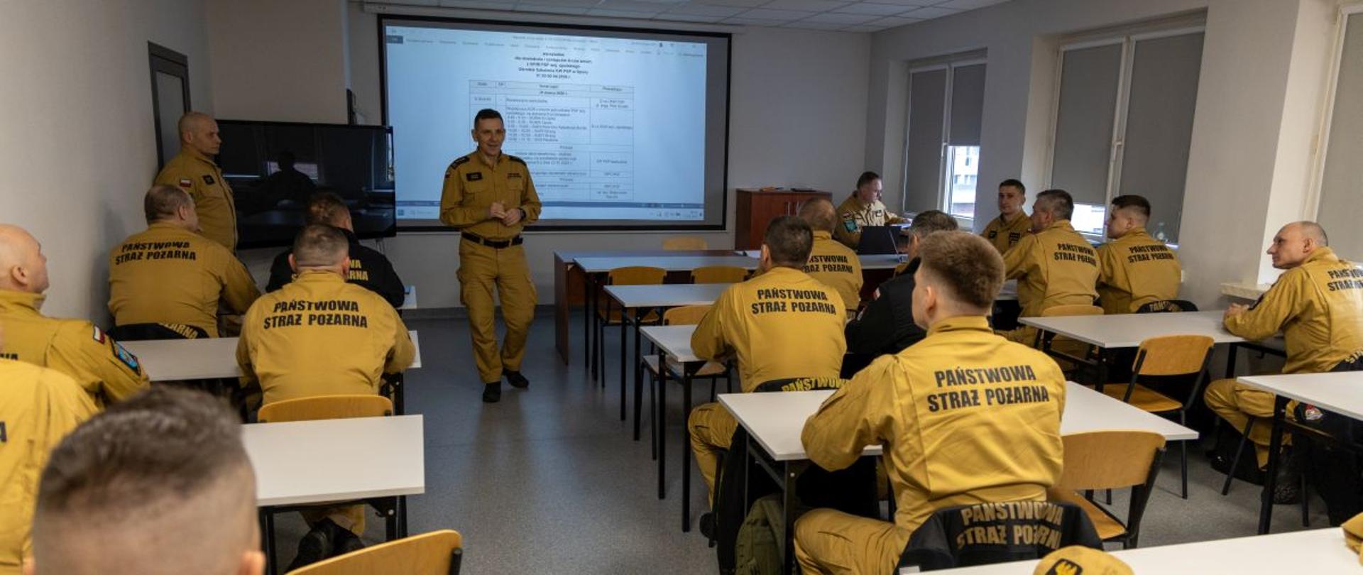 Grupa funkcjonariuszy PSP w żółtych koszulkach z napisem „Państwowa Straż Pożarna” siedzi w sali wykładowej podczas warsztatów dowódców zmian. Uczestnicy słuchają wystąpienia Zastępcy Opolskiego Komendanta Wojewódzkiego PSP. Na ekranie wyświetlony jest dokument. W pierwszym rzędzie znajduje się stanowisko komputerowe prowadzącego.
