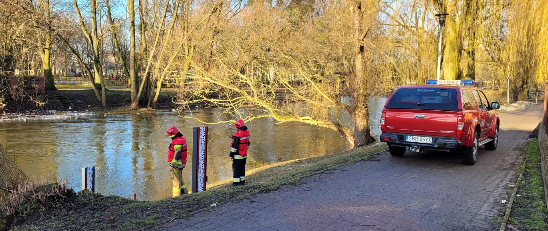 Zdjęcie przedstawia dwóch strażaków odczytujących stan wody na wodowskazie na rzece Drwęca. Z prawej strony samochód operacyjny brodnickiej Komendy.