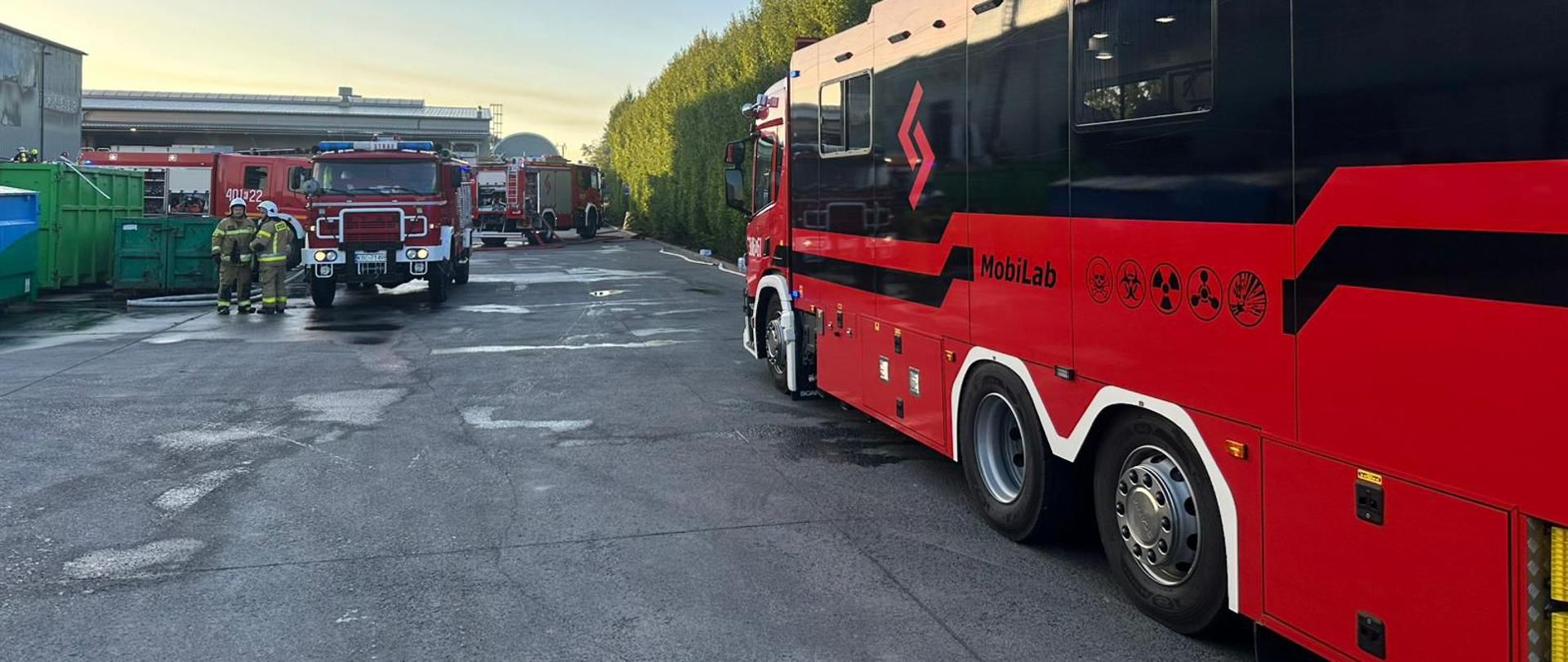 Na terenie zakładu znajduje się kilka strażackich samochodów. Nie widać żadnego dymu ani ognia. Dwóch strażaków rozmawia ze sobą. Strażacy ubrani w ubrania specjalne oraz hełmy.