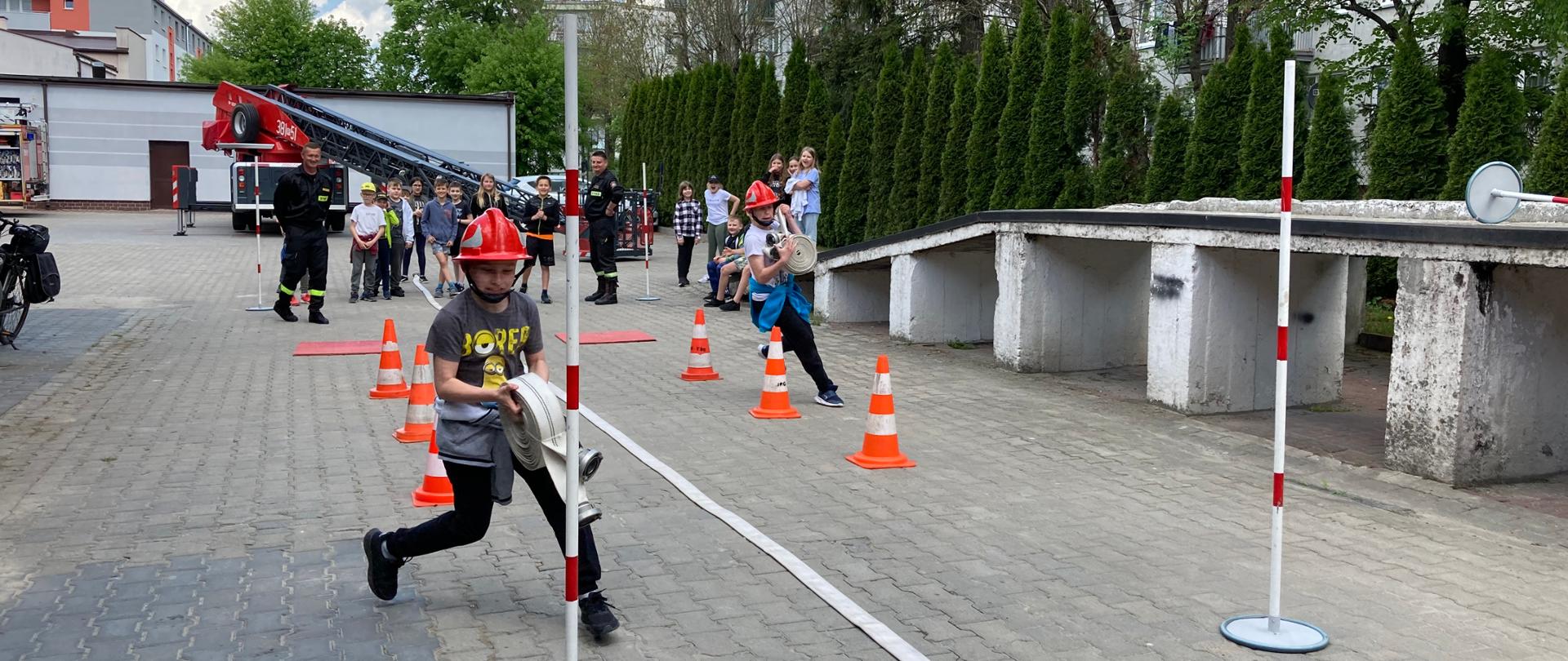 Na placu strażnicy ustawiony tor przeszkód z pachołkami. Tor pokonuje dwoje dzieci, które biegną, trzymając w ręku węże. 