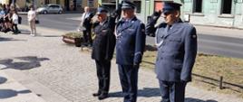 Plac przed Pomnikiem Żołnierza Polskiego w Rawiczu. Delegacja rawickich służb mundurowych (PSP, Policji i SW) oddaje honor poprzez salutowanie, po wczesniejszym złożeniu wiązanki kwiatów pod tablicą pamiętkową. W tle kamienice.