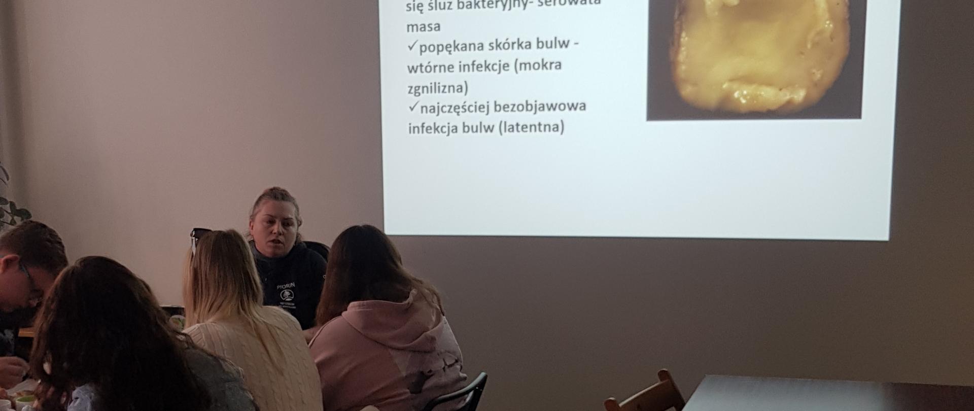 Sala szkoleniowa, na której siedzą uczestnicy spotkania oraz prowadząca. Na ścianie wyświetlony slajd ze zdjęciem choroby ziemniaka.