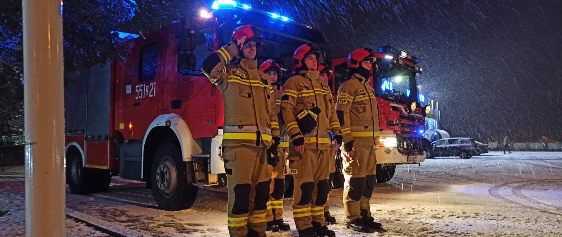 Zdjęcie przedstawia strażaków ustawionych w dwuszeregu przed dwoma samochodami strażackim z włączonymi światłami mijania oraz niebieskimi błyskowymi. Jeden ze strażaków salutuję. Zdjęcie wykonano nocną porą. Podczas zdjęcia występowały opady śniegu.