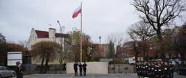 Poczet flagowy podnosi flagę na maszt przed nimi stoją strażacy w dwóch rzędach oraz przed nimi strażak oddaje honor.