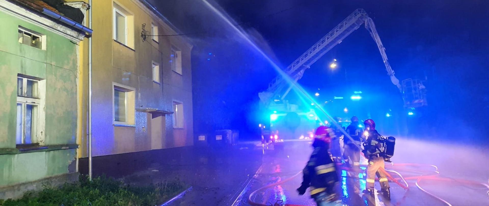 Zdjęcie przedstawia miejsce pożaru w porze nocnej. Budynek trzykondygnacyjny wielorodzinny w którym płonie dach. Strażacy z rozstawionym podnośnikiem przygotowują stanowisko gaśnicze, strażacy w aparatach podają prądy wody na palący się dach, jeden ze strażaków wynosi butlę 11 kilogramową z gazem propan-butan. 