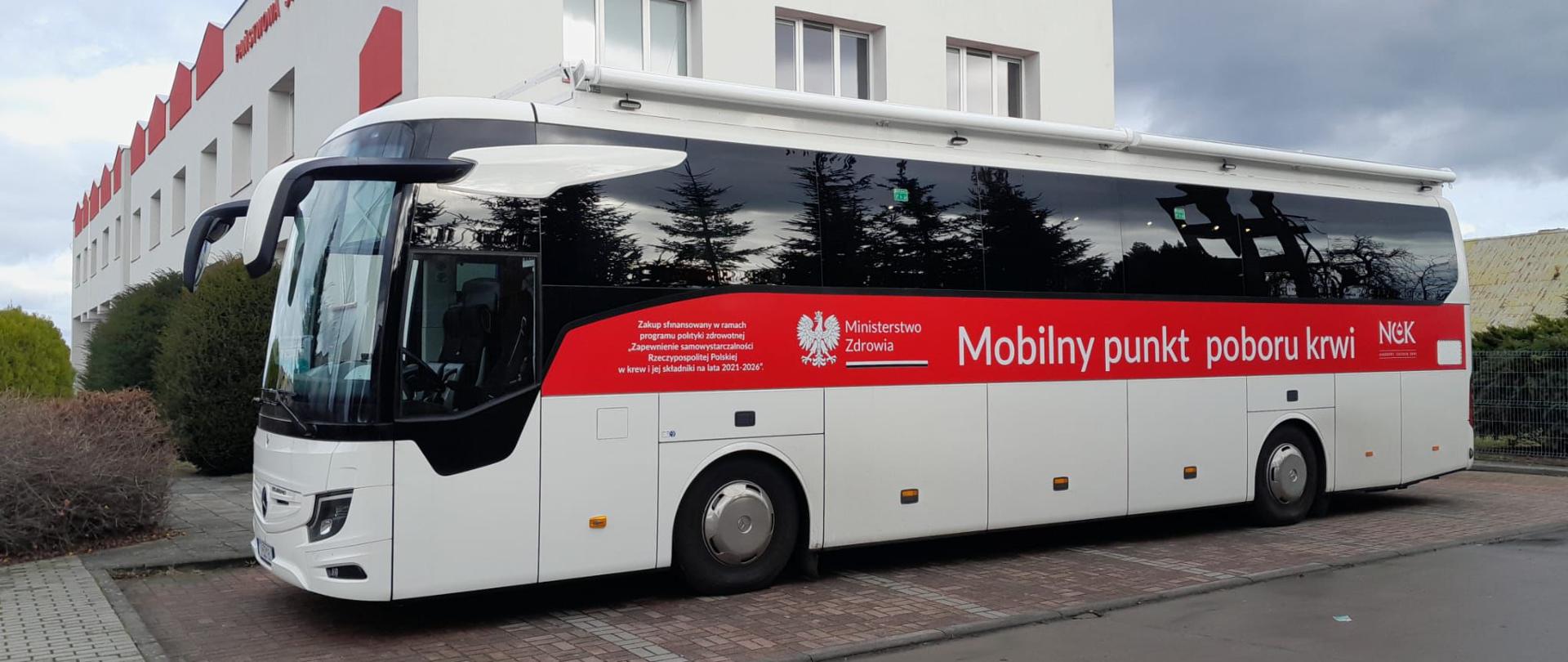 Zdjęcie przedstawia autobus "Mobilny punkt poboru krwi", który stoi na parkingu na tle budynku Komendy Powiatowej Państwowej Straży Pożarnej.