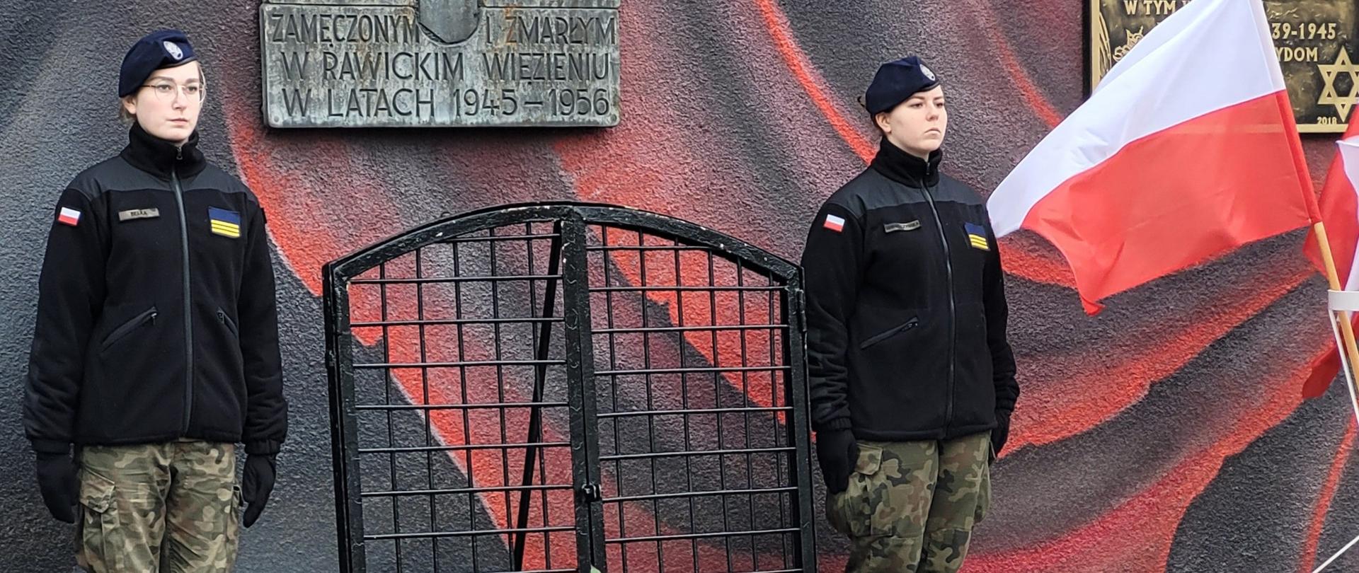 Mur więzienny a na nim zawieszone sa tablice pamiątkowe. Przed tablicami, na chodniku leżą wiązanki kwiatów. Na warcie stoją dwie uczennice klasy mundurowej. Z prawej strony powiewa flaga biało-czerwona. 