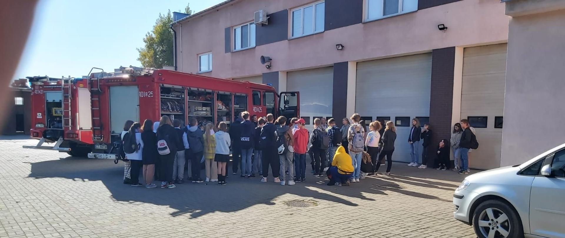 1. Zdjęcie przedstawia dwie grupy uczniów siódmych klas szkoły podstawowej wraz z nauczycielkami w liczbie 63 osoby , stojące obok dwóch wozów strażackich przy których znajduje się zmiana służbowa w liczbie 5 osób pełniąca służbę tego dnia. Strażacy oraz uczniowie i nauczycielki stoją na szarej kostce przed garażami przy wozach strażackich. Strażacy ubrani są na czarno w strój koszarowy oraz posiadają na głowach czapki z logiem państwowej straży pożarnej. Za nimi stoją dwa wozy strażackie koloru czerwonego pierwszo oraz drugo wyjazdowy z otwartymi skrytkami na sprzęt strażacki. W tle widnieją bramy garażu jednostki ratowniczo-gaśniczej w Aleksandrowie kujawskim koloru białego ,elewacja jest koloru jasno różowego z wstawkami z płytek klinkierowych koloru brązowego.