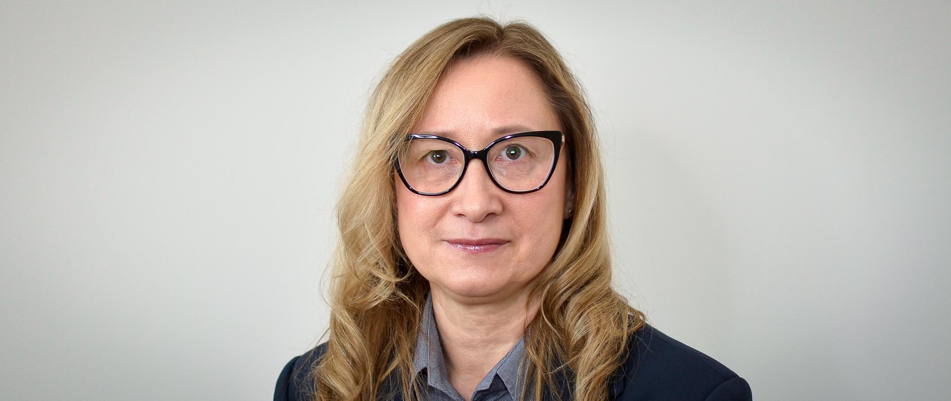 Kobieta w ciemnej marynarce, jaśniejszej bluzce, okularach, z jasnymi, długimi włosami