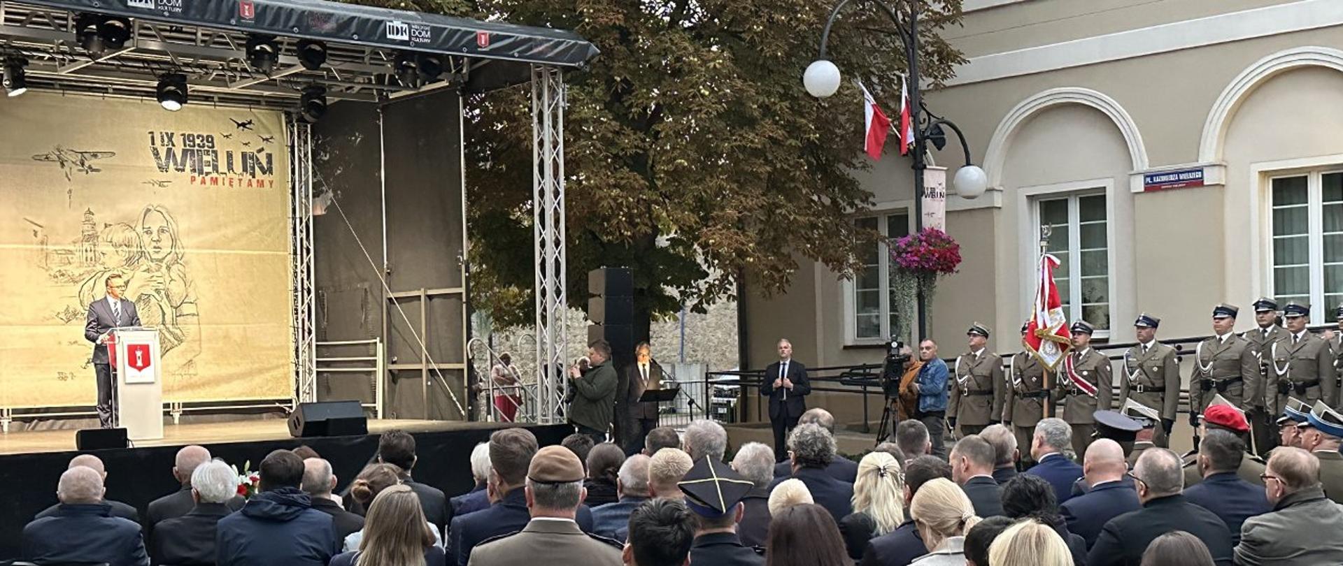 Zdjęcie przedstawia dużą publiczność, która siedzi na krzesłach i patrzy na scenę, na której mężczyzna przemawia do mikrofonu. Na dużej płachcie w tle widnieje napis "1 IX 1939 WIELUŃ PAMIĘTAMY". Z boku sceny stoi kilku żołnierzy.