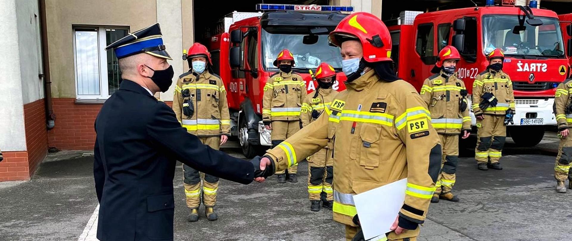 Zdjęcie przedstawia Komendanta Powiatowego Państwowej Straży Pożarnej w Będzinie mł. bryg. Arkadiusz Spera podczas wręczania decyzji objęcia stanowiska Dowódcy Jednostki Ratowniczo-Gaśniczej w Będzinie przez mł. bryg. Krzysztofa Roka. Zdjęcie wykonano na palcu Komendy. W tle widać strażaków w ubraniach specjalnych oraz samochody pożarnicze.