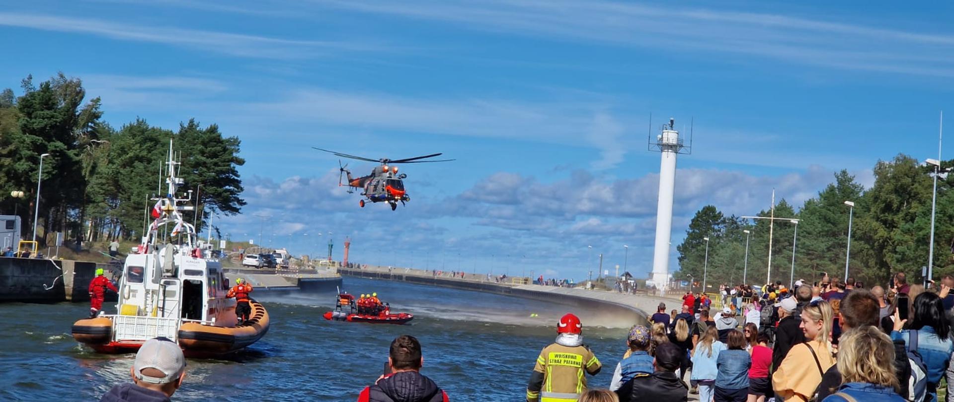Zdjęcie przedstawia pokazy ratownicze Morskiej Służby Poszukiwania i Ratownictwa w kanale łebskim przez śmigłowiec, statek ratowniczy i ponton ratowniczy.