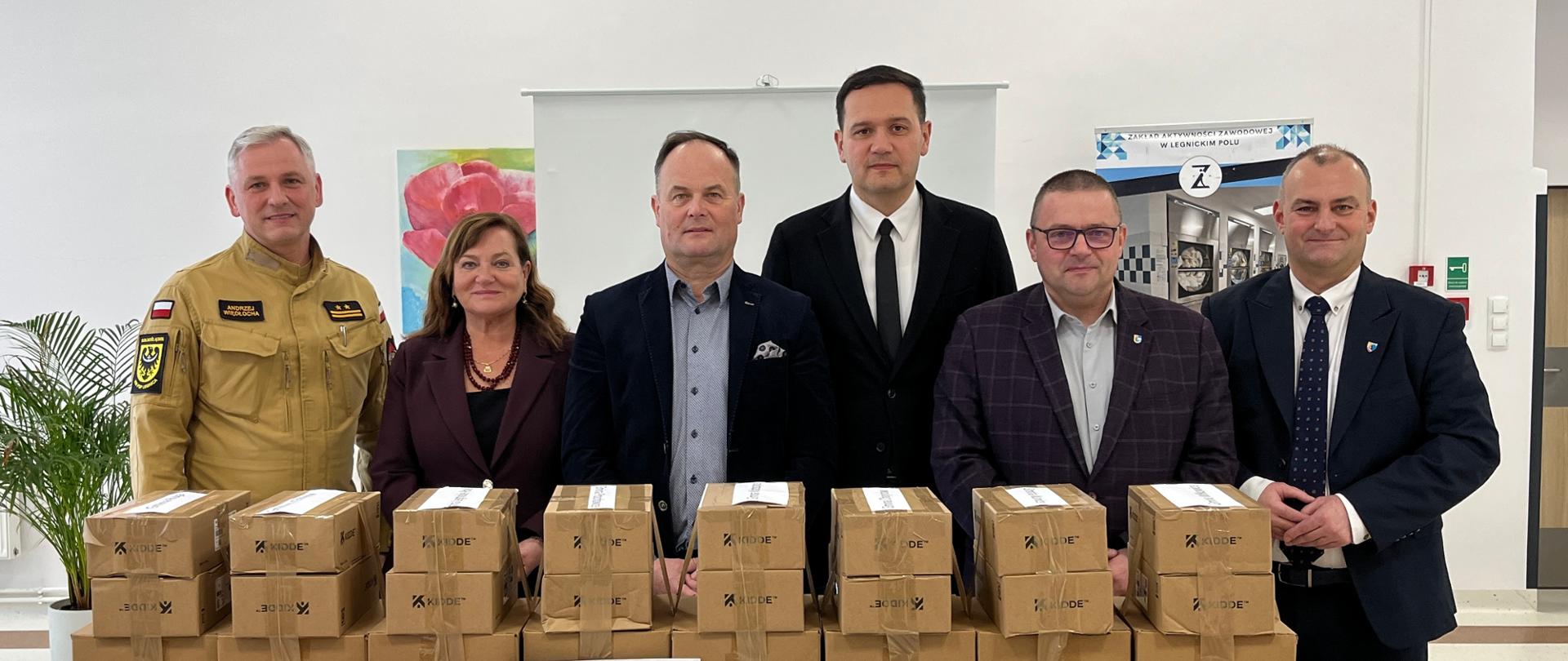 Zdjęcie przedstawia grupę sześciu osób stojących w rzędzie za stołem w jasnym pomieszczeniu o białych ścianach. W skład grupy wchodzi pięciu mężczyzn i jedna kobieta. Po lewej stronie stoi mężczyzna w piaskowym mundurze strażackim z widocznymi naszywkami, obok niego kobieta w bordowym żakiecie i naszyjniku, a dalej czterej mężczyźni w strojach formalnych – garniturach i marynarkach. Na stole przed nimi ustawione są liczne tekturowe pudełka ułożone w dwóch rzędach, a przed nimi wyeksponowano mniejsze opakowania z czujnikami dymu i czadu oraz czerwoną tabliczkę z napisem „Czujka na straży Twojego bezpieczeństwa”. W tle, za uczestnikami, widać biały ekran projekcyjny, kolorowy obraz na ścianie oraz roll-up promujący lokalną instytucję. Po lewej stronie kadru widoczna jest zielona roślina doniczkowa, co ociepla oficjalny charakter fotografii dokumentującej przekazanie sprzętu bezpieczeństwa.
