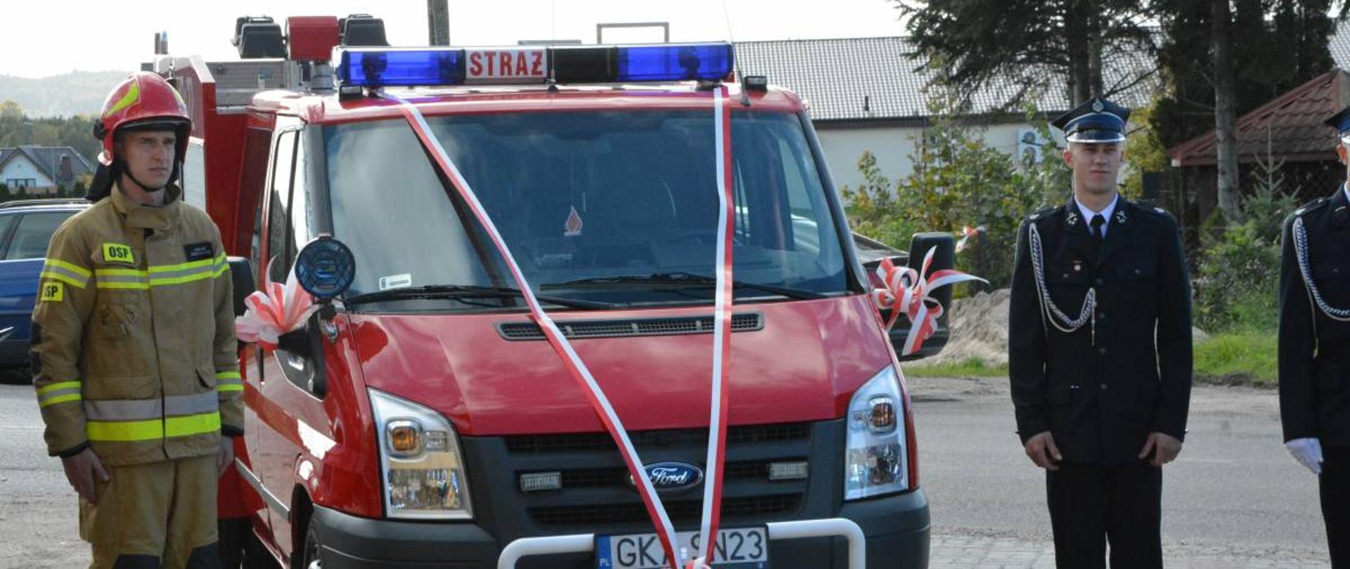 Przyozdobiony biało czerwonymi wstążkami samochód pożarniczy, obok którego stoją strażacy.