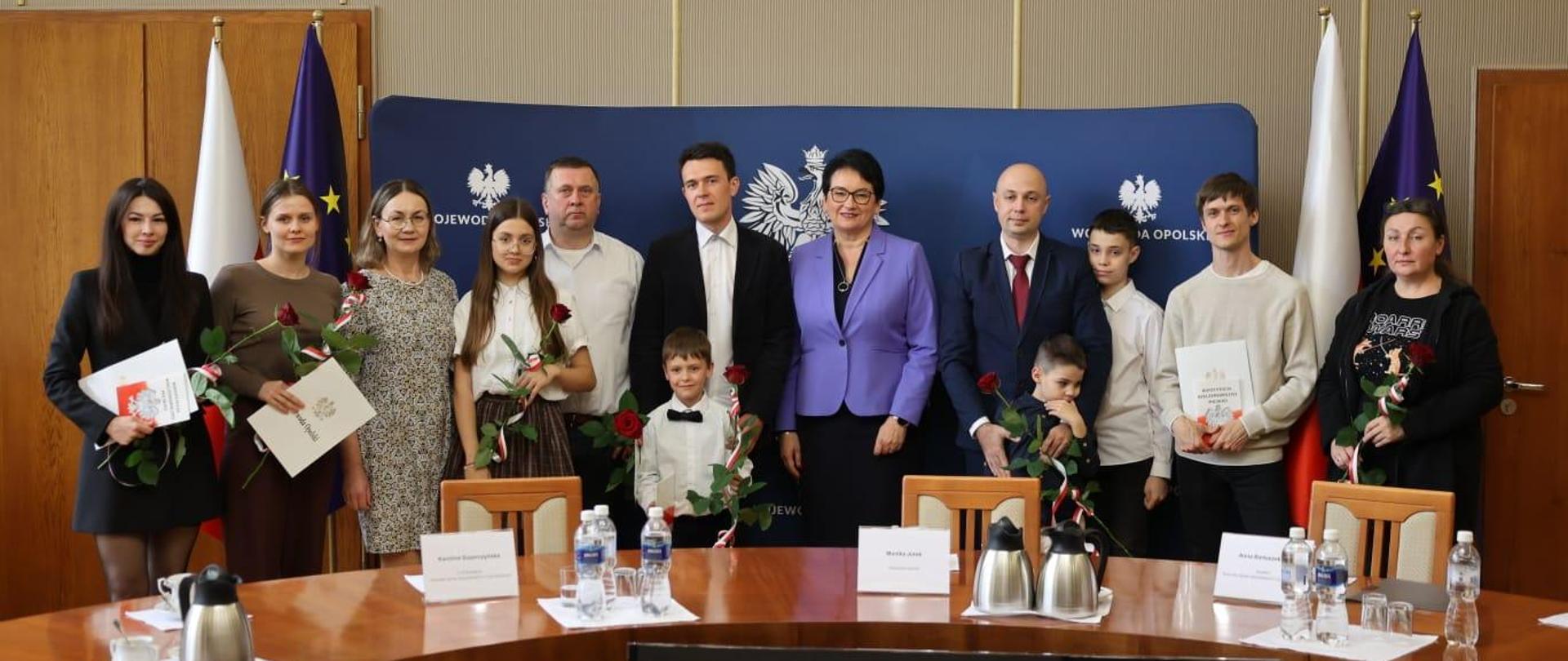 Na zdjęciu widać uczestników - nowych, polskich obywateli, którzy podczas uroczystości w Opolskim Urzędzie Wojewódzkim odebrali akty obywatelstwa polskiego. Wszyscy stoją obok siebie i pozują do zdjęcia, część z nich ma w rękach róże oraz akty. Na środku stoi Wojewoda Opolski. 