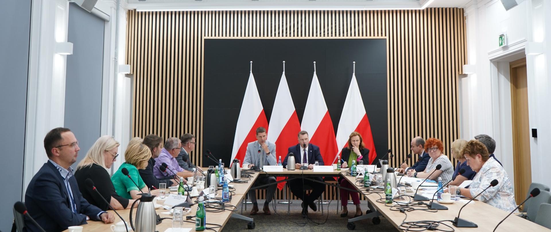 Widok na salę, przy ustawionych w czworokąt stołach siedzą ludzie, u szczytu stołu minister Czarnek, wiceminister Piontkowski i kobieta w czarnej marynarce.