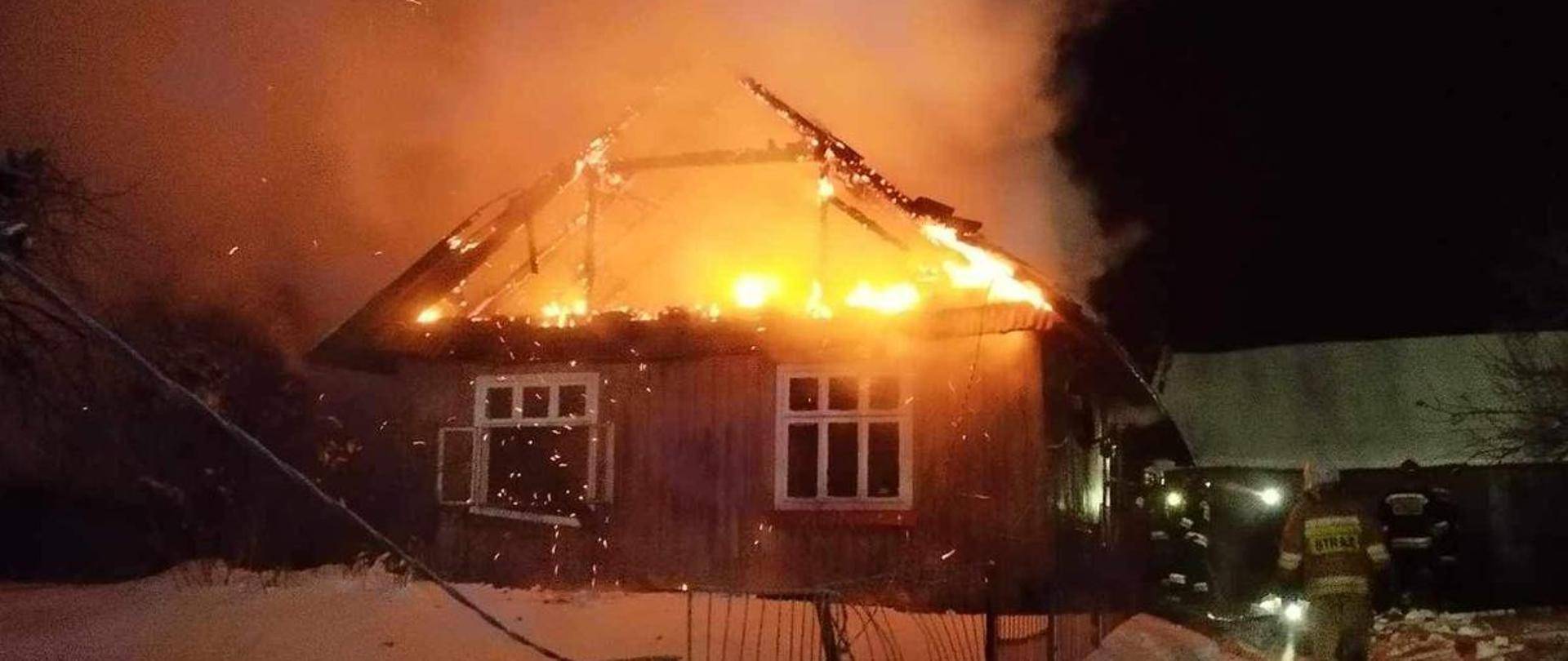 Na zdjęciu widoczny jest palący się drewniany budynek jednorodzinny, z którego unosi się dym. Z prawej strony widoczni są pracujący strażacy, którzy ubrani są w ubranie specjalne. Za nimi widoczny jest budynek gospodarczy.