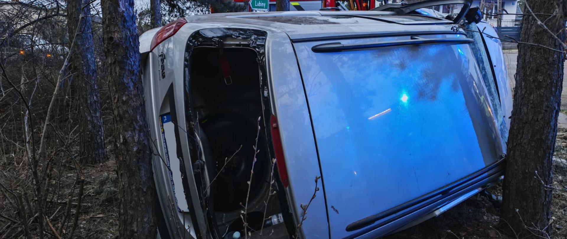 Samochód osobowy leży na boku między drzewami, poza jezdnią, po wypadku. Jedno z okien jest rozbite, a wnętrze pojazdu częściowo widoczne. W tle widać wóz strażacki i zabudowania mieszkalne, co sugeruje trwającą akcję ratunkową. Scena ma miejsce o zmierzchu lub przy słabym oświetleniu, z niebieskimi światłami odbijającymi się na karoserii.