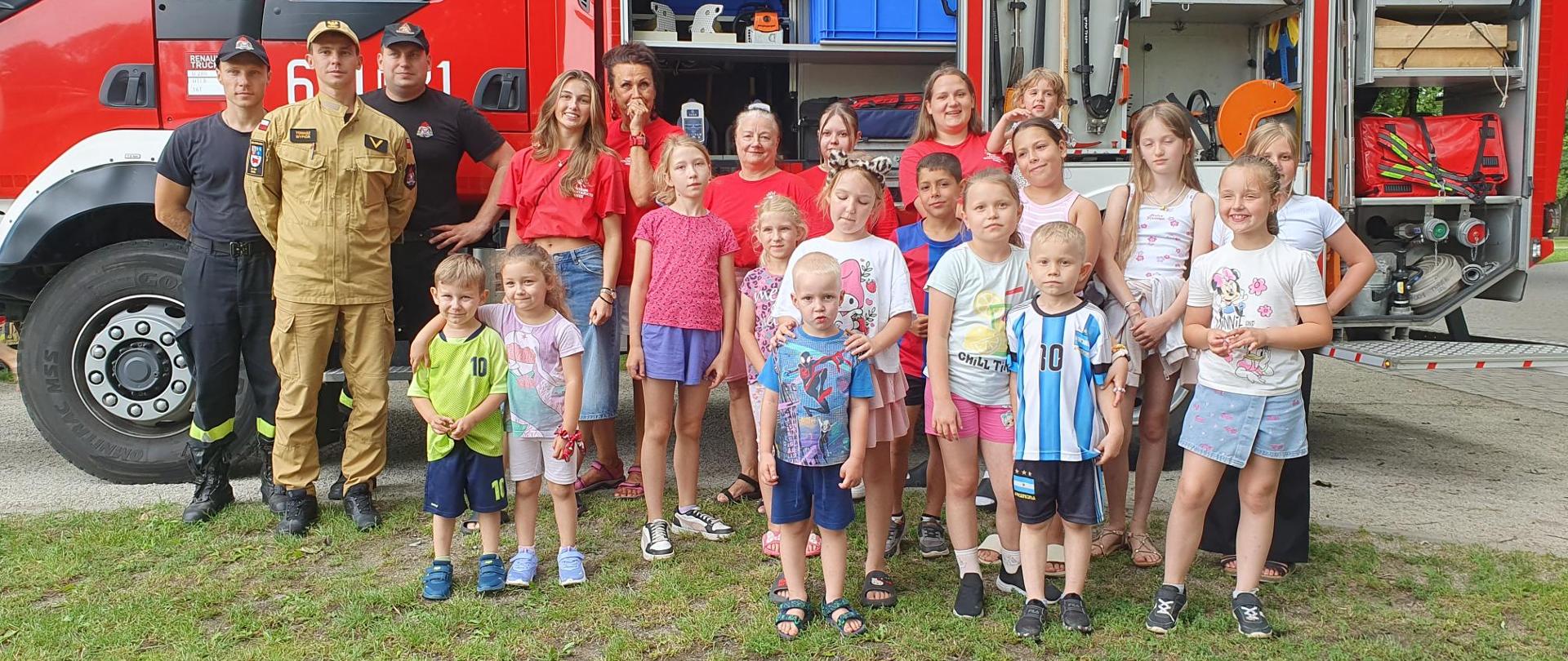 Pamiątkowe zdjęcie grupowe uczestników spotkania, wolontariuszy oraz strażaków, pozujących na tle czerwonego wozu strażackiego z otwartymi skrytkami ze sprzętem. Wszyscy uśmiechają się w stronę obiektywu, a dzieci stoją w pierwszym planie.