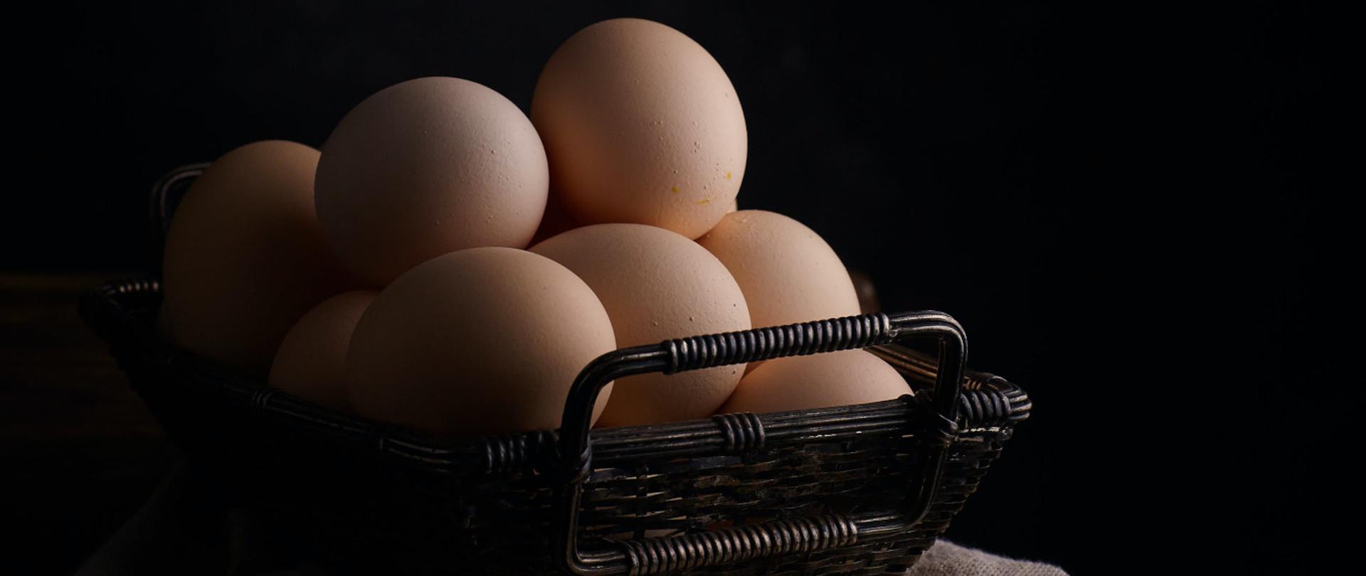 Ciemno brązowy koszyk z jajkami stojący na stoliku przykrytym szarym obrusem