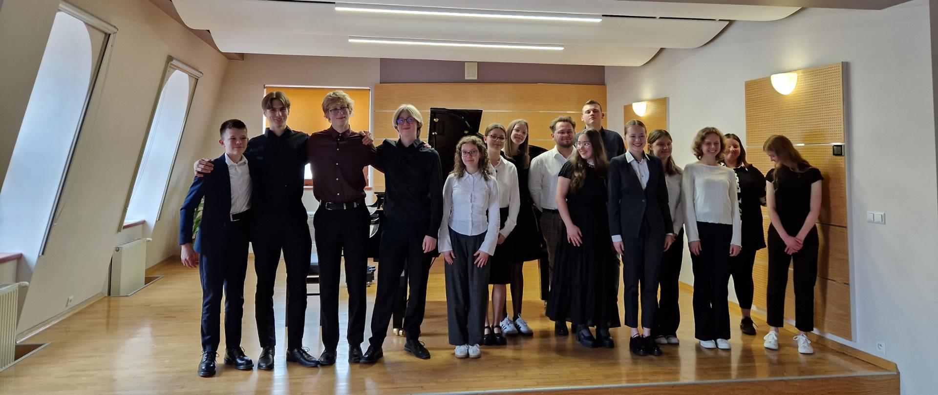 Zdjęcie przedstawia grupę około piętnastu młodych ludzi, prawdopodobnie nastolatków, stojących na drewnianej scenie w przestronnej auli. Większość z nich ubrana jest w eleganckie stroje: chłopcy w garnitury, a dziewczęta w ciemne sukienki lub białe koszule i ciemne spodnie/spódnice. Stoją w luźnych grupach, niektórzy uśmiechają się, a niektórzy mają bardziej poważne miny. Scena jest podwyższona, a za nimi widać beżowe ściany i ukośne sufity z wbudowanymi lampami. Po prawej stronie zdjęcia widoczne są również kinkiety ścienne, rzucające ciepłe światło. Pomieszczenie wydaje się być dobrze oświetlone. Na lewo od sceny widać białe ściany z oknami w kształcie trapezu. Ogólnie zdjęcie emanuje atmosferą uroczystego wydarzenia, być może koncertu lub występu.